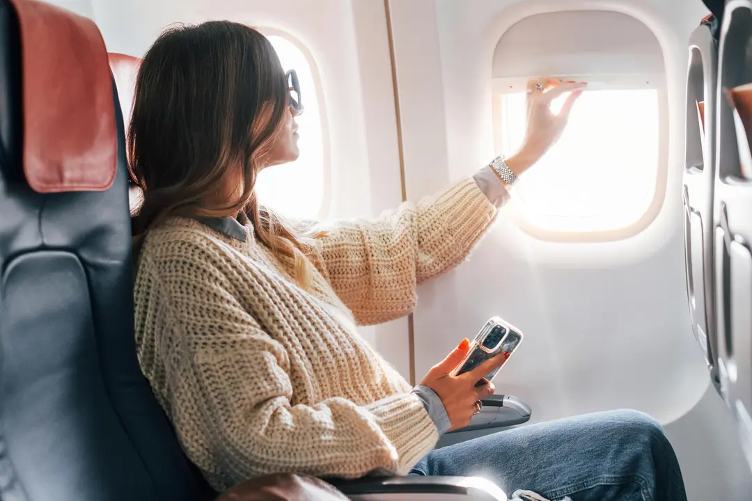 Sitting with phone in hands. Young female passanger in casual clothes is in the plane.