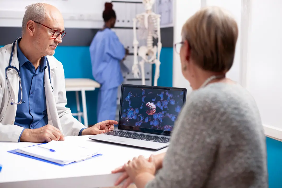 Senior male physician in lab coat discusses virus image on laptop with retired woman in well equipped hospital room. Old doctor and female patient looking at enlarged viral cells on digital device.