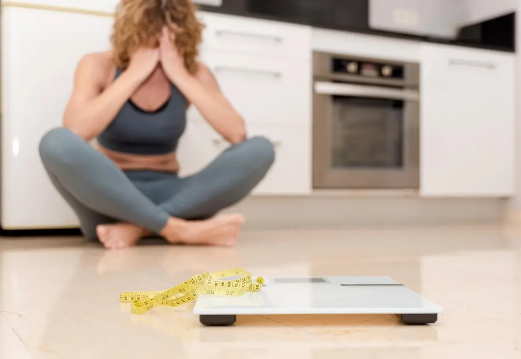 Upset woman in sportswear sitting on kitchen floor with scale and measuring tape, weight loss struggle
