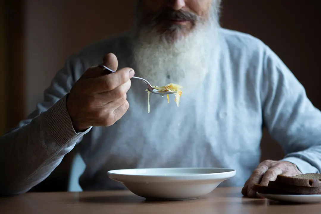 Old sad man with a long gray beard sitting by the table and eating soup and bread