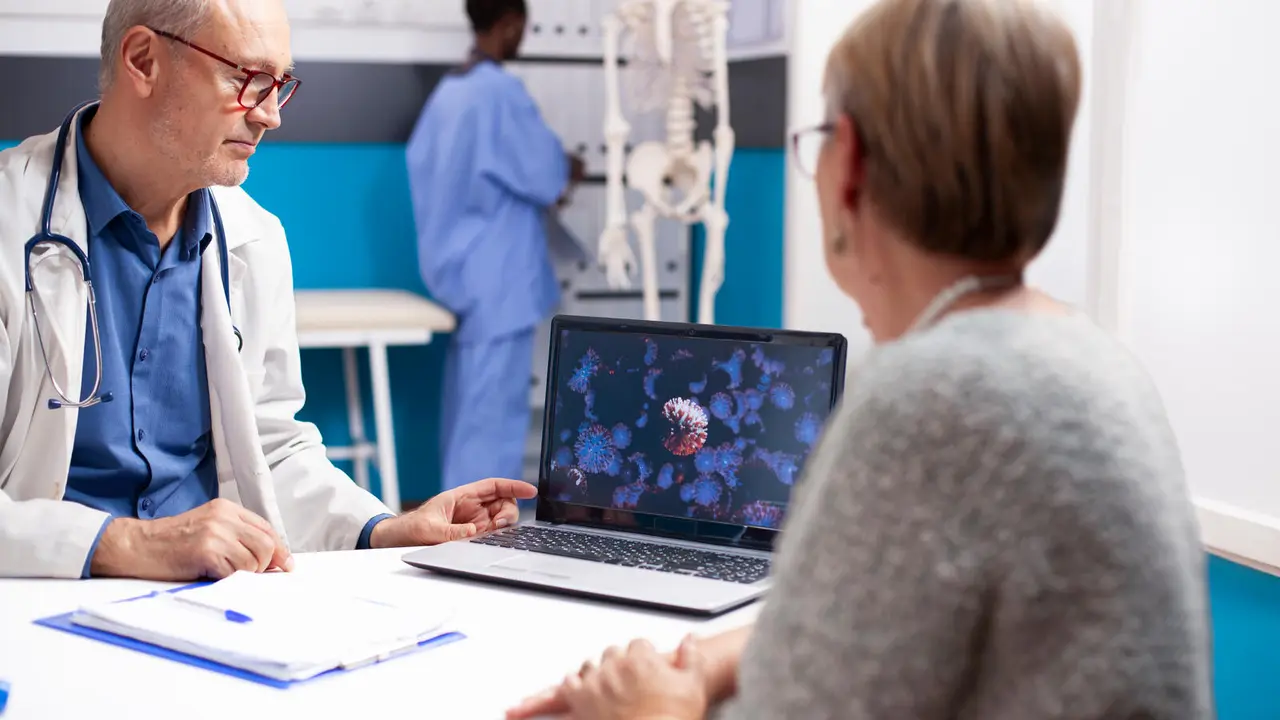 Senior male physician in lab coat discusses virus image on laptop with retired woman in well equipped hospital room. Old doctor and female patient looking at enlarged viral cells on digital device.