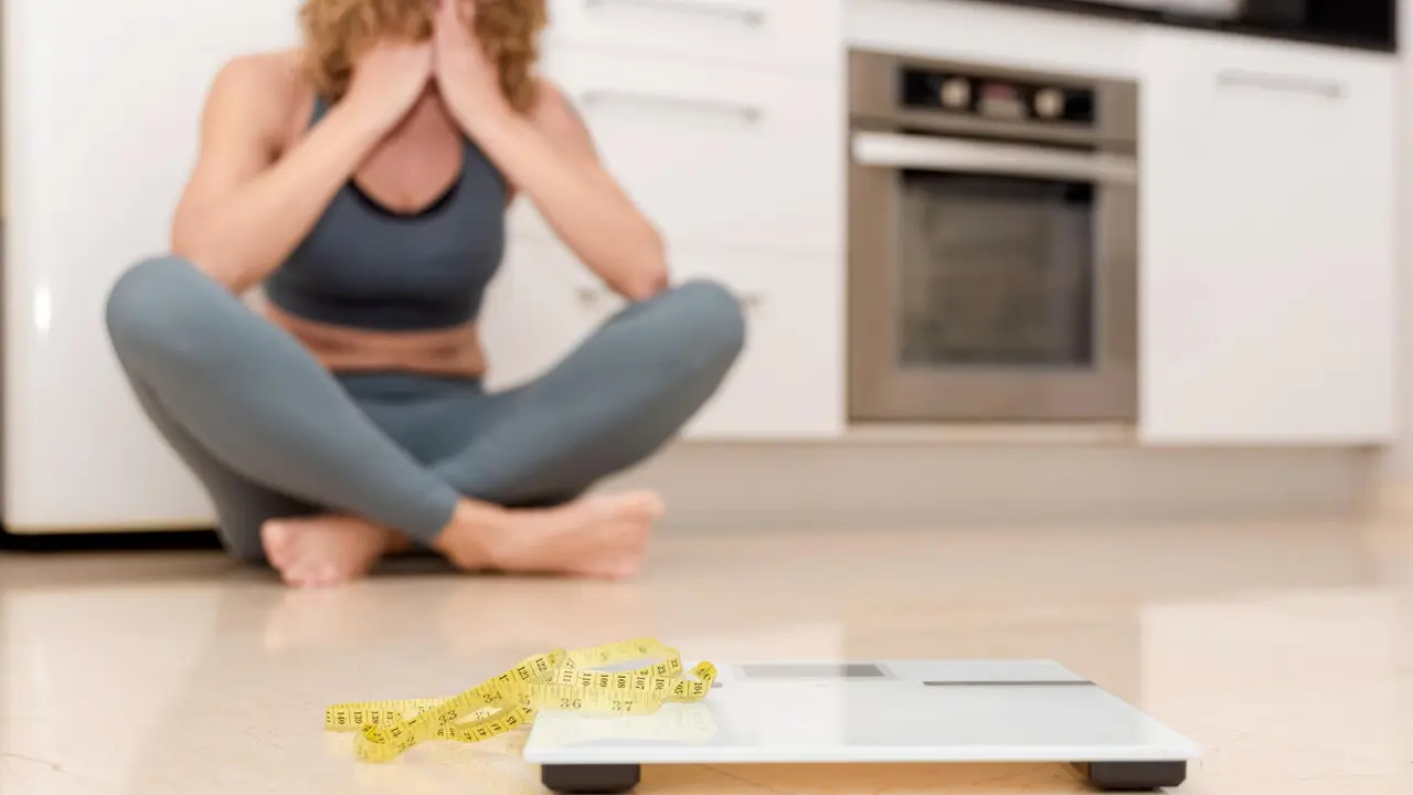 Upset woman in sportswear sitting on kitchen floor with scale and measuring tape, weight loss struggle