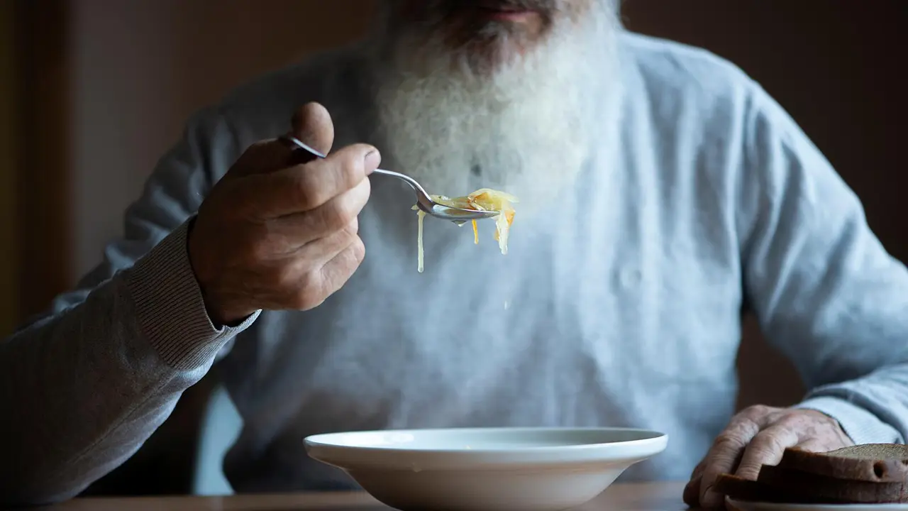 Old sad man with a long gray beard sitting by the table and eating soup and bread